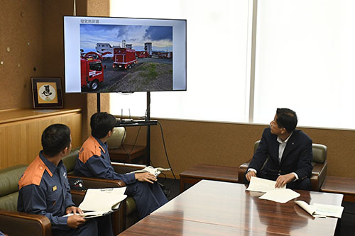 活動中の写真を見ながら説明を聞く江原市長(右)
